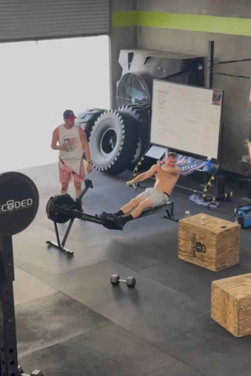 A member rowing while another walks by with a tractor tire on Freedom WOD night